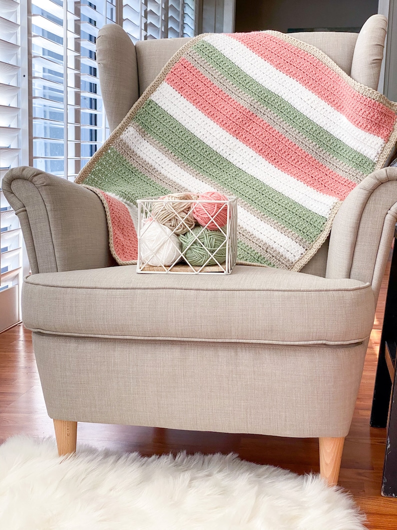 Op de afbeelding: Een gebreid gestreept deken in tinten roze, groen, wit en beige, gedrapeerd over een beige fauteuil met houten poten. Een draadmand gevuld met bollen garen in bijpassende kleuren staat op de armleuning.