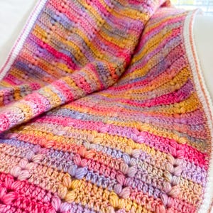 May include: A colorful crocheted blanket with a striped pattern in shades of pink, purple, yellow, and orange. The blanket is made with a textured stitch that creates a raised, bumpy surface.