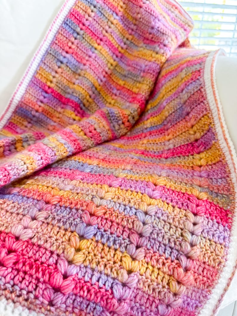 May include: A colorful crocheted blanket with a striped pattern in shades of pink, purple, yellow, and orange. The blanket is made with a textured stitch that creates a raised, bumpy surface.