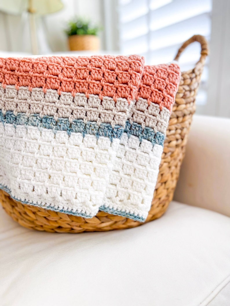 May include: A crocheted blanket with stripes of coral, beige, blue and white. The blanket is folded and placed in a wicker basket.