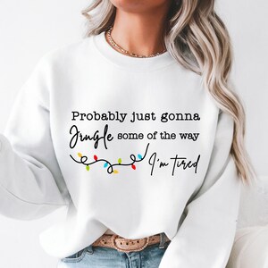 May include: White sweatshirt with the text "Probably just gonna jingle some of the way, I'm tired" in black script. A string of colorful Christmas lights is below the text. The sweatshirt is a casual, festive garment.