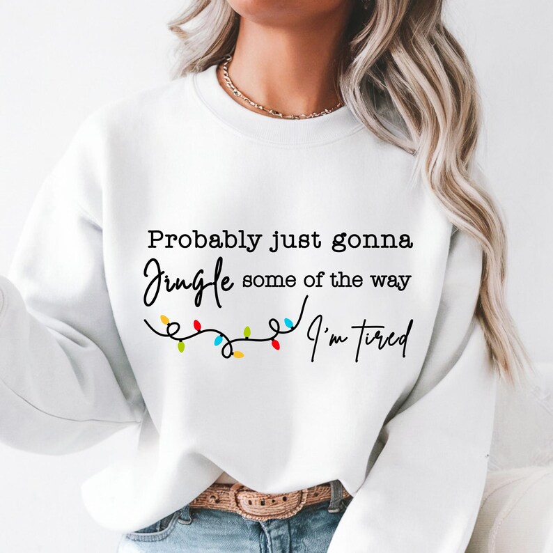 May include: White sweatshirt with the text "Probably just gonna jingle some of the way, I'm tired" in black script. A string of colorful Christmas lights is below the text. The sweatshirt is a casual, festive garment.