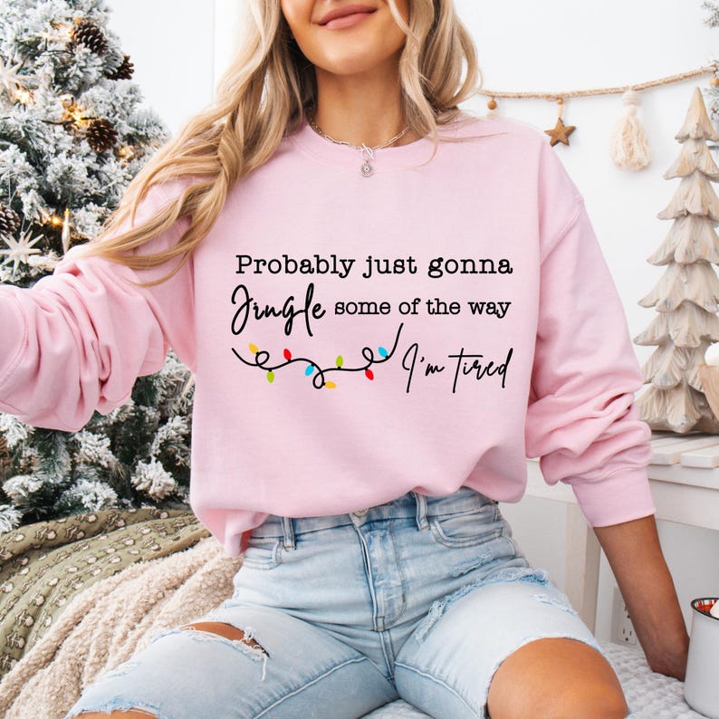 May include: A light pink sweatshirt with the text "Probably just gonna Jingle some of the way I'm tired" in black script. A string of colorful Christmas lights is below the text. The sweatshirt is a crew neck style.