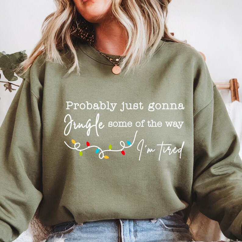 May include: Olive green sweatshirt with the text "Probably just gonna jingle some of the way, I'm tired" in white script. A string of colorful Christmas lights is below the text. The sweatshirt is a casual, comfortable style.