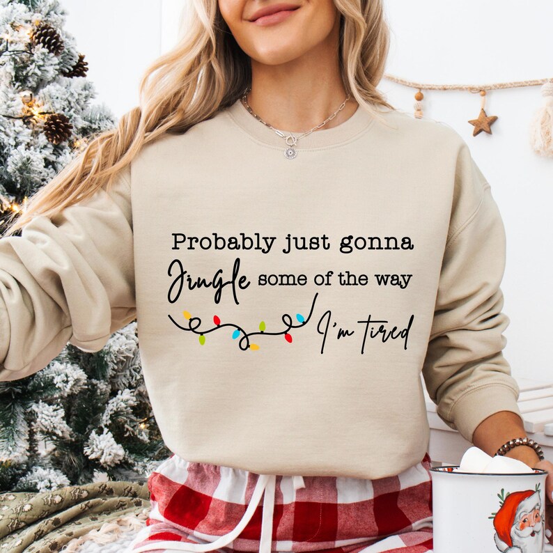 May include: Beige sweatshirt with the text "Probably just gonna jingle some of the way, I'm tired" in black script. A string of colorful Christmas lights is below the text. The sweatshirt is a neutral color.