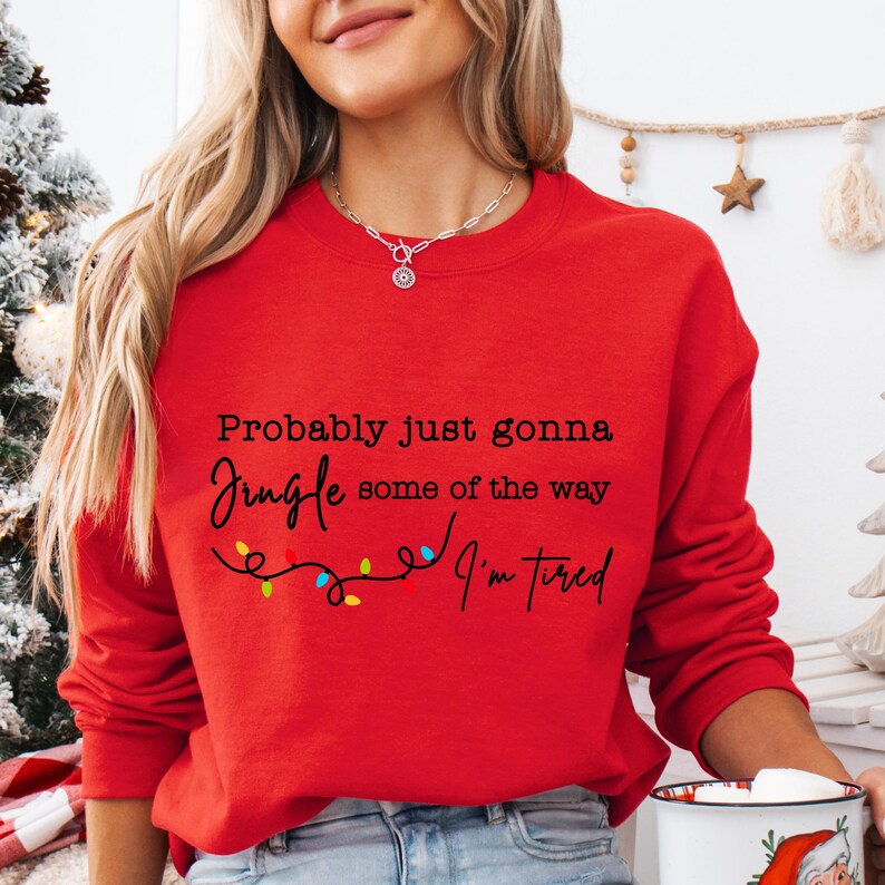 May include: A red sweatshirt with the text "Probably just gonna jingle some of the way, I'm tired" in black script. A string of colorful Christmas lights is below the text. The sweatshirt is a crew neck style.