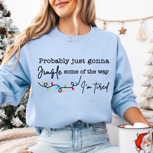 May include: Light blue sweatshirt with the text "Probably just gonna Jingle some of the way I'm tired" in black script. A string of colorful Christmas lights is below the text. The sweatshirt is a crew neck style.