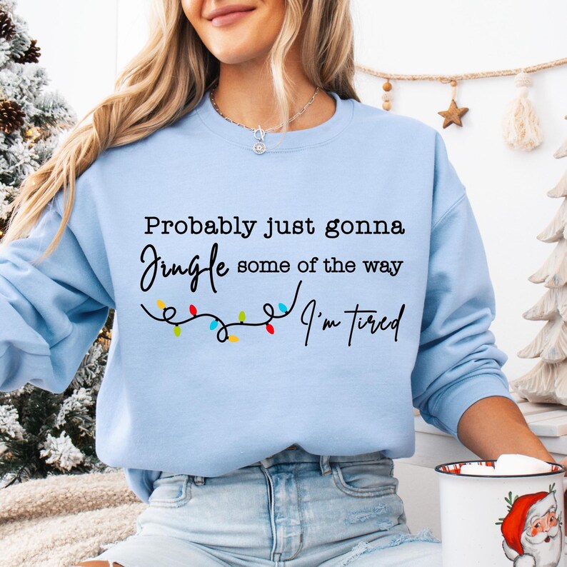 May include: Light blue sweatshirt with the text "Probably just gonna Jingle some of the way I'm tired" in black script. A string of colorful Christmas lights is below the text. The sweatshirt is a crew neck style.