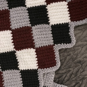 May include: A close-up of a crocheted blanket with a checkered pattern in shades of gray, white, black, and brown. The blanket has a scalloped edge.