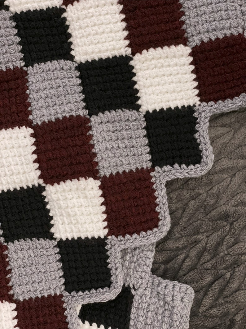 May include: A close-up of a crocheted blanket with a checkered pattern in shades of gray, white, black, and brown. The blanket has a scalloped edge.