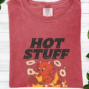 Puede incluir: Camiseta de color carmesí con las palabras "HOT STUFF" en una fuente audaz y desgastada. Un diablo de dibujos animados con un tridente aparece debajo del texto. La camiseta es de la marca Comfort Colors.