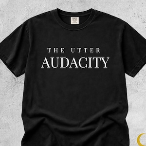 Może przedstawiać: Czarny t-shirt z białym napisem "THE UTTER AUDACITY" pośrodku. Koszulka ma okrągły dekolt i krótkie rękawy. Nazwa marki "LUNA DRIP" znajduje się w prawym dolnym rogu.