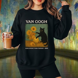 Puede incluir: Sudadera negra con un diseño de "Girasoles" de Van Gogh y un gato negro. El diseño incluye el texto "VAN GOGH" encima de la imagen y "SUNFLOWERS (THIRD VERSION) - 1888" debajo.