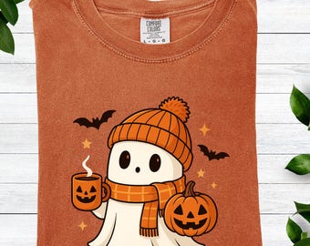 Puede incluir: Una camiseta naranja Comfort Colors con un fantasma de dibujos animados que lleva un gorro y una bufanda naranjas, sosteniendo una taza de calabaza y de pie junto a una calabaza tallada. El diseño también incluye murciélagos y estrellas.