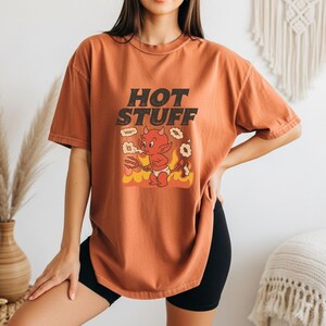Könnte beinhalten: Rostfarbenes T-Shirt mit dem Text "HOT STUFF" über einer Cartoon-Teufel-Grafik. Der Teufel ist rot mit einem Dreizack und Flammen. Das Shirt ist kurzärmlig und hat eine lockere Passform.