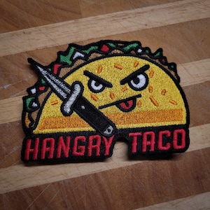 Può includere: Una toppa ricamata con un taco di cartone animato con un'espressione arrabbiata che tiene un coltello. Il taco ha una lingua rossa e un topping di lattuga verde e rossa. Il testo "Hangry Taco" è ricamato in rosso su uno sfondo nero.