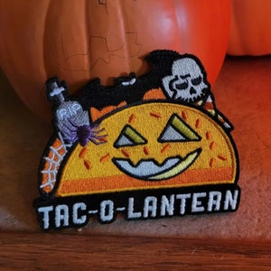 May include: Embroidered patch with a jack-o'-lantern taco design, featuring a smiling face, a bat, and a skull. The patch includes the text "TAC-O-LANTERN" and a tombstone with "RIP". The patch is orange, yellow, black, and white.