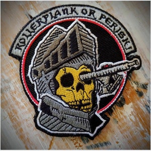 Puede incluir: Un parche bordado en blanco y negro con una calavera que lleva un casco y una pistola apuntando a su ojo. El texto "TOWERFLANK OR PERISH" está alrededor del exterior del parche.