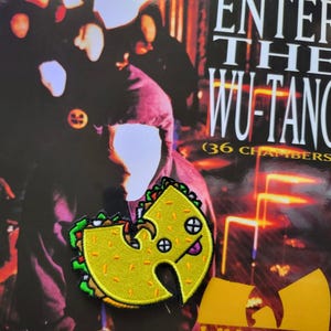 Pode incluir: Um remendo bordado amarelo do logotipo do Wu-Tang Clan, em forma de símbolo do Wu-Tang Clan, com um rosto sorridente e a língua de fora. O remendo está em cima de uma capa de álbum do Wu-Tang Clan, que diz "Enter the Wu-Tang (36 Chambers)".