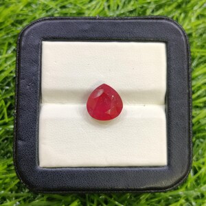Puede incluir: Un rubí facetado en forma de pera, exhibido en una caja de joyería negra y blanca. El rubí es de un color rojo vibrante y parece estar bien pulido. La caja se encuentra sobre un fondo de hierba verde.