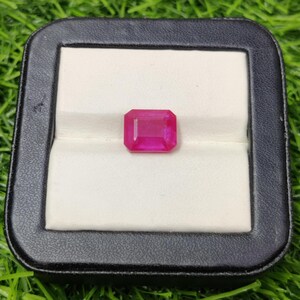 Puede incluir: Un rubí de corte rectangular, de un tono rosa intenso, presentado en una caja de joyería negra con interior blanco. Las facetas de la gema capturan la luz, resaltando su claridad. La caja se encuentra sobre un fondo de hierba verde.