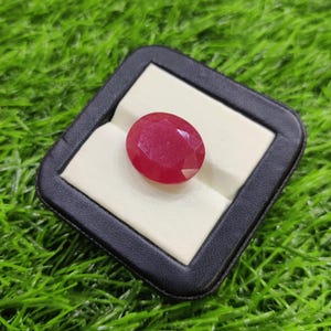 Puede incluir: Una gema de rubí ovalada y facetada, presentada en una caja de joyería de polipiel negra con interior blanco. El rubí es de color rojo intenso y descansa sobre tela blanca. El fondo es verde.