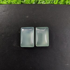 May include: Two rectangular, pale green gemstones with rounded corners are displayed. The stones have a translucent quality, reflecting light. The gems are set against a dark background, highlighting their color and shape. These could be used for jewelry.