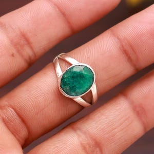 Puede incluir: Un anillo de plata con una gran esmeralda ovalada. La esmeralda es de color verde vibrante y está engastada en una banda de caña dividida. El anillo se muestra en una mano.