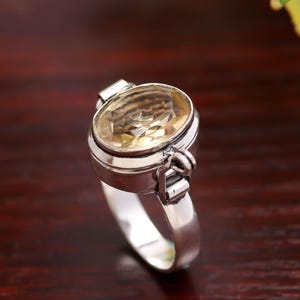 May include: A silver ring with an oval, pale yellow gemstone. The gemstone is set in a bezel setting with decorative side details. The ring is shown on a dark wooden surface, with yellow flowers in the background.