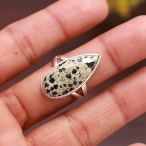 May include: A silver ring with a teardrop-shaped stone. The stone has a speckled pattern of black spots on a cream background. The ring's band is a simple, thin silver design.