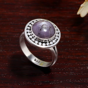 Peut inclure: Bague en argent avec une grosse pierre précieuse ronde et violette. La pierre est entourée d'une lunette de petites perles d'argent. L'anneau de la bague est lisse et poli. Le numéro "925" est estampillé sur l'anneau.