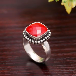 Puede incluir: Anillo de plata con una gema roja cuadrada y facetada. La gema está rodeada por un borde de cuentas. El anillo está sobre un fondo de madera oscura, con flores amarillas en la esquina superior derecha.