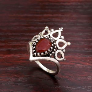 May include: A silver ring featuring a teardrop-shaped, deep red gemstone. The ring has a crown-like design around the stone, with openwork details. The band is smooth and polished. A piece of jewellery.