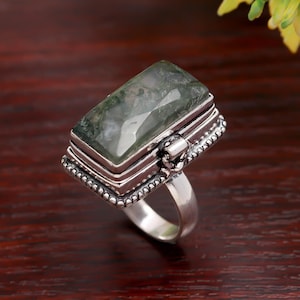 May include: A rectangular silver ring featuring a large, moss-green gemstone. The gemstone is set in a detailed, layered bezel setting with a beaded border. The ring's band is a simple, polished silver.