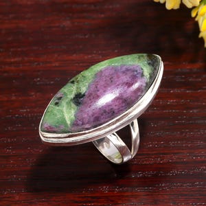 May include: A silver ring with a large, oval gemstone. The gemstone displays a striking mix of green, purple, and black colours. The ring's band is a simple, polished silver design.