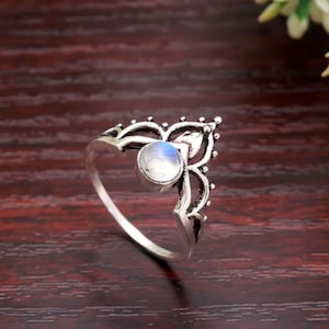 Könnte beinhalten: Silberner Ring mit einem runden, schillernden Mondstein, der in einem verzierten, durchbrochenen Design gefasst ist. Der Ring hat ein dekoratives, stilisiertes Blumen- oder Blattformmotiv auf dem Band und um den Stein. Auf dunkler Holzoberfläche.