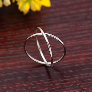 May include: A silver ring with a distinctive crisscross design. The ring has two intersecting bands forming an X shape. It is set against a dark wood surface, with yellow flowers visible in the background.