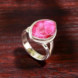May include: A sterling silver ring featuring a large, oval-shaped, vibrant pink gemstone. The gemstone is set in a bezel setting, and the ring has a split shank design. The ring is displayed on a dark wood surface.