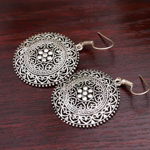 May include: A pair of silver-colored, round earrings with intricate, openwork designs. Each earring features a central floral pattern surrounded by detailed scrollwork and a beaded edge. The earrings have a hook closure.