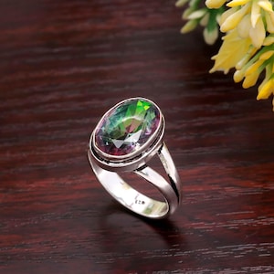 Può includere: Un anello in argento con una gemma ovale che mostra uno spettro di colori, tra cui verde, viola e giallo. L'anello ha un design a gambo diviso e una finitura lucida, che mette in risalto la brillantezza della gemma. Gioielli.