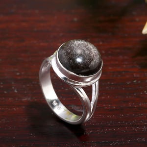 May include: A silver ring with a large, round, black gemstone. The gemstone has a speckled appearance. The ring's band is split near the gemstone, creating a unique design. The ring is set against a dark wood surface.