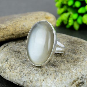 May include: A silver ring with a large, oval, off-white gemstone. The ring is set on a textured gray stone, with green foliage in the background. The gemstone has a smooth, polished surface.