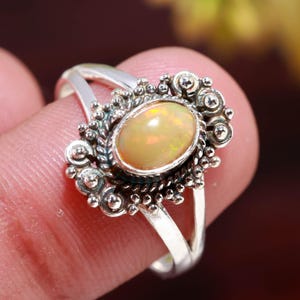 May include: A silver ring featuring an oval, light-yellow opal gemstone. The opal is set in a decorative, ornate setting with a detailed, textured design. The ring's band splits into two at the top, adding to its unique style.