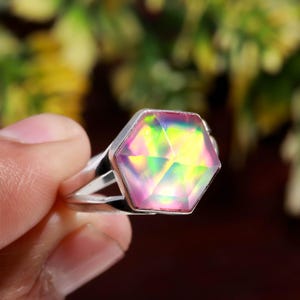 May include: A silver ring featuring a large, iridescent, hexagonal gemstone. The gemstone displays a vibrant array of colors, including pink, yellow, and green. The ring's band splits into two parallel lines on either side of the gemstone.