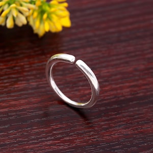 Op de afbeelding: Een zilverkleurige open ring met een glad, afgerond ontwerp. De ring heeft een opening aan de bovenkant, waardoor een open stijl ontstaat. Gele bloemen staan op de achtergrond, tegen een donkere houten ondergrond. Sieraden.
