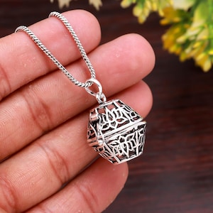 May include: Silver pendant necklace with a geometric cage design. The pendant hangs from a fine chain. The intricate openwork pattern of the pendant is visible, showcasing the detailed craftsmanship. The necklace is held in a hand.