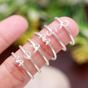 May include: A collection of silver-toned initial rings, each featuring a different letter of the alphabet. The rings are displayed on a finger, showcasing their delicate design and polished finish. The letters are in a simple, elegant font.