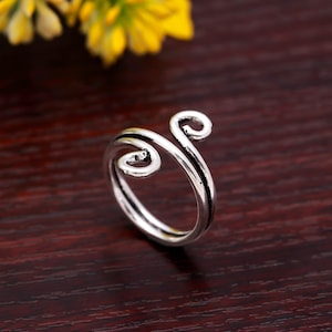 May include: A silver-colored ring with a unique, swirling design. The ring features two curved, spiral-shaped elements at the top, connected by a band. The ring is set against a dark wooden surface, with yellow flowers in the background.