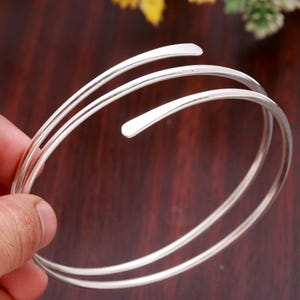 May include: A silver-colored metal bangle bracelet featuring a multi-coil wrap design. The bracelet consists of two overlapping circular bands with smooth, rounded ends that taper slightly, creating a continuous spiral appearance in a polished finish.
