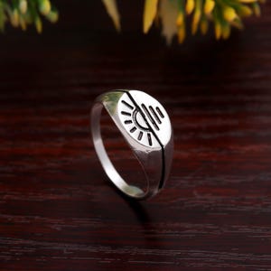 May include: A silver ring with a sunburst design on a rounded face. The ring has a split design with a black line down the center. The ring is on a dark wood surface.